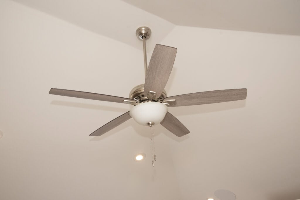 A modern ceiling fan with five wooden blades and a central light fixture is mounted on a white ceiling. Two pull chains hang from the light. Recessed lights are also visible on the ceiling.