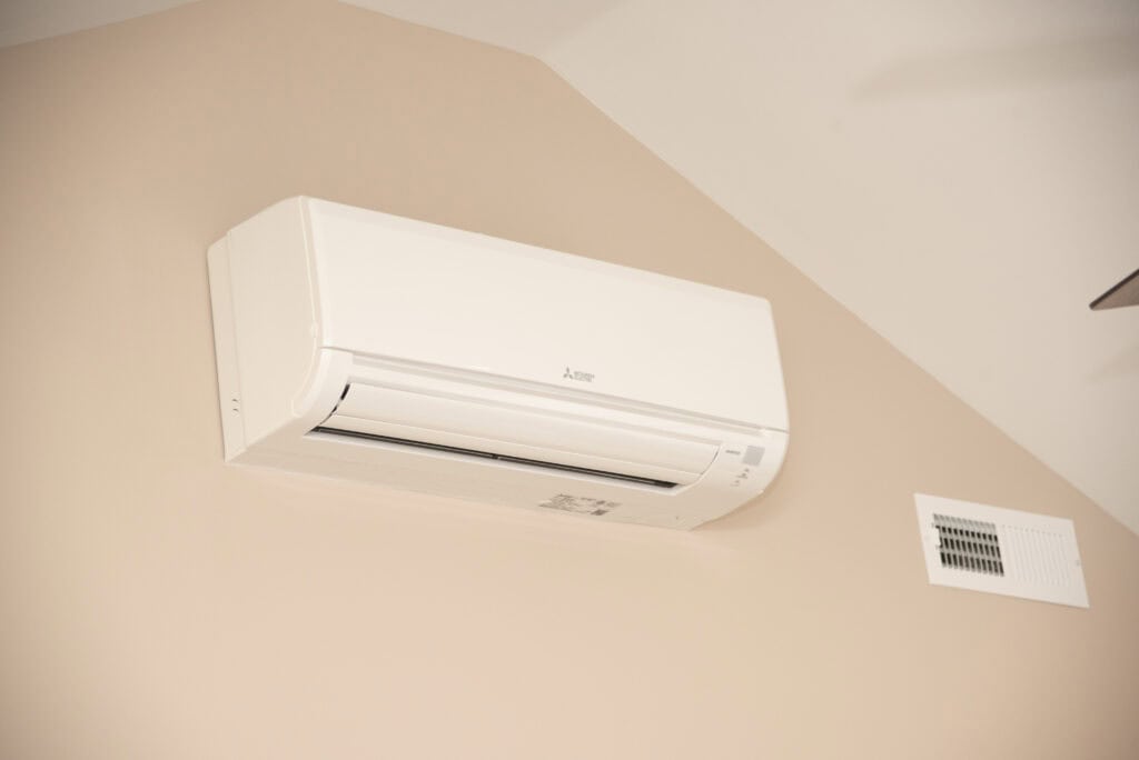 A white wall-mounted air conditioning unit is installed on a beige wall above an air vent. The unit is rectangular and compact, designed for cooling or heating the room.