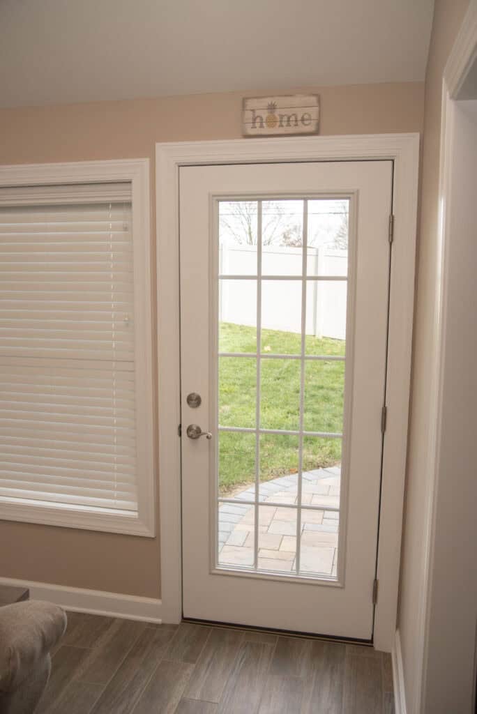 A glass-paneled white door leads to a backyard with green grass and a stone walkway. A window with closed blinds is on the left, and a small home sign hangs above the door.