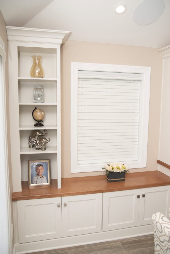 A built-in white shelving unit displays decor items, a globe, a framed photo, and an elephant figurine. A window with closed blinds is above a wooden bench with white cabinets and a small flower arrangement.