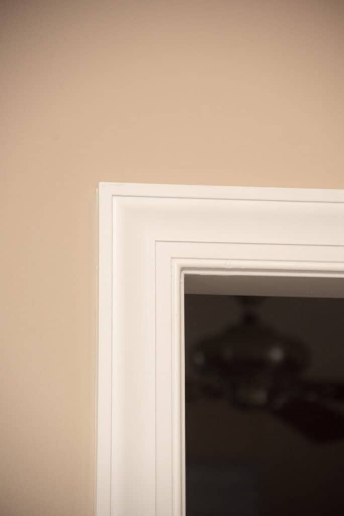Close-up of a white-framed mirror or window against a beige wall, with the reflection of a ceiling fan partially visible in the glass.