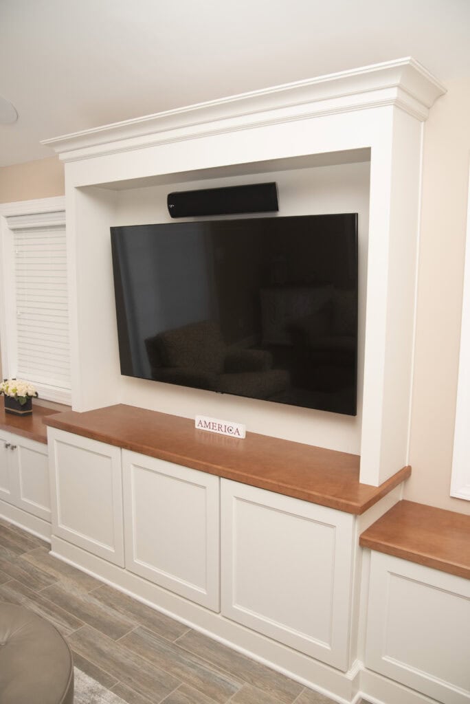 A flat-screen TV is mounted on a white built-in entertainment unit with wood countertops. Below the TV is a small sign that says “AMERICA.” There’s a soundbar above the TV and a window with closed blinds on the left.