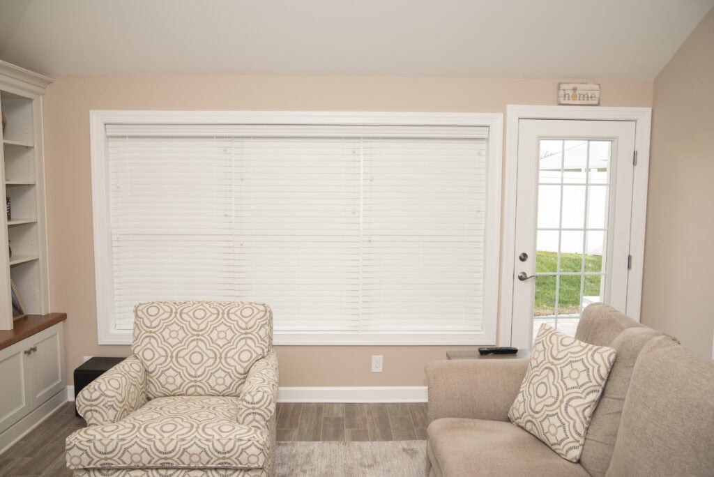 A cozy living room with two beige armchairs, one with a patterned cushion, facing a large window with white blinds. A glass door to the right leads outside, and built-in shelves are partially visible on the left.