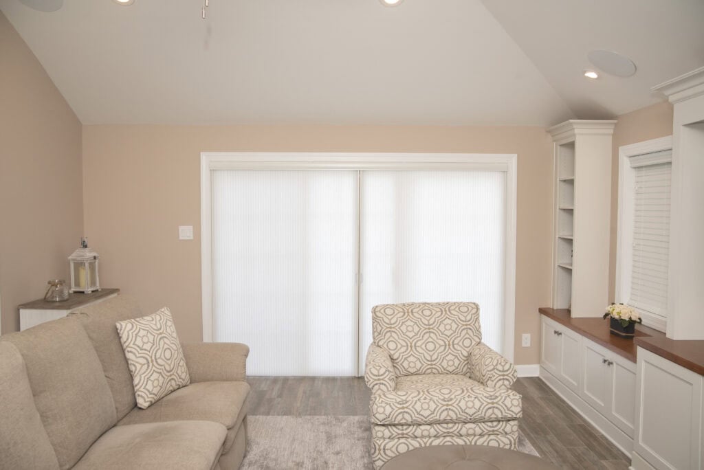 A bright living room with beige walls, a light-patterned armchair, a beige sofa, built-in white shelves, wood accents, and large sliding doors covered by sheer white curtains.