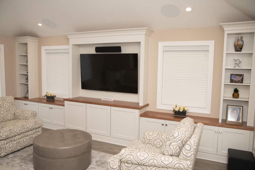 A cozy living room features a wall-mounted TV between two shuttered windows, surrounded by built-in white shelves and cabinets. Two patterned armchairs and a round ottoman sit in front. Decorative items and flowers are on the shelves.