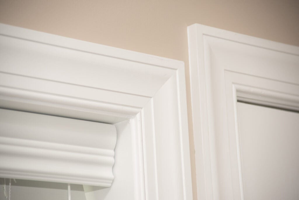 Close-up of two white door or window frames with molding, set against a beige wall. The frames have clean lines and sharp corners, highlighting the details of the trim work.