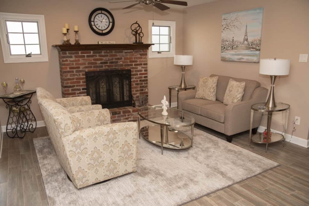 A cozy living room with a brick fireplace, beige sofa and armchairs, a glass coffee table, lamps, wall art of the Eiffel Tower, and wooden floors with a light rug. Two windows let in natural light.