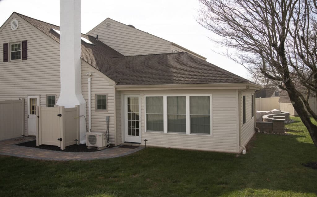 A beige house with a small extension featuring large windows, a white chimney, outdoor HVAC unit, and a neatly maintained backyard with grass and a leafless tree.