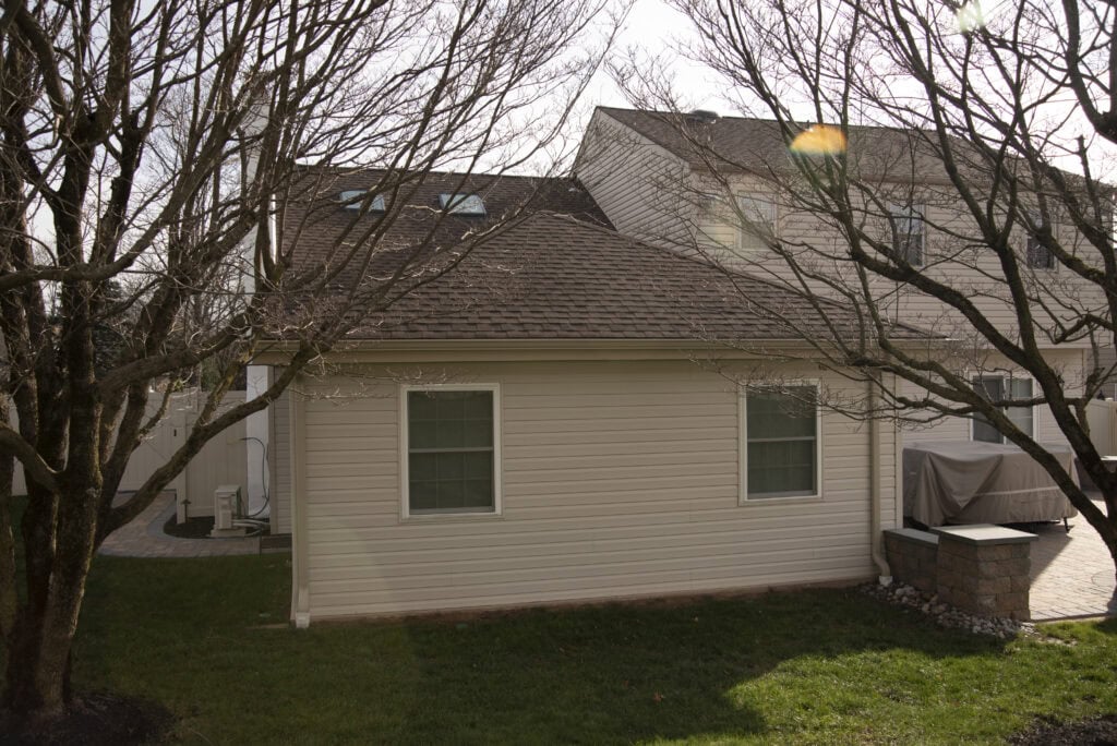 A beige detached garage or small house with two windows, surrounded by leafless trees and a green lawn, on a sunny day. A covered grill and stone patio are visible to the right.