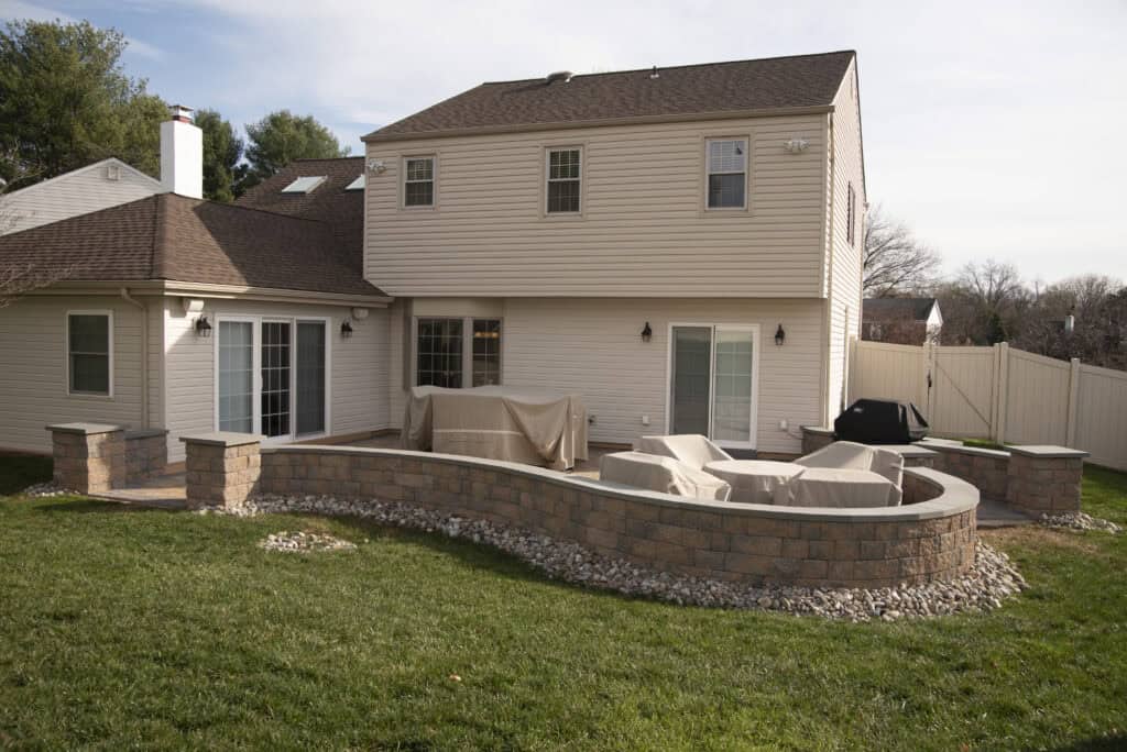 A suburban backyard features a two-story beige house with a stone patio, low curved stone wall, covered patio furniture, and a grill, surrounded by a green lawn and white privacy fence.