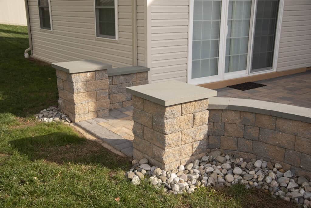 A stone patio with pillars and a curved low wall, bordered by white decorative rocks, next to a house with beige siding and large sliding glass doors.