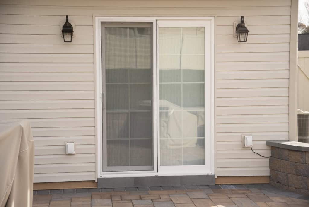 A closed white sliding glass patio door with a screen, set in a beige siding wall. Two black wall lanterns flank the door, and a brick patio extends in front.