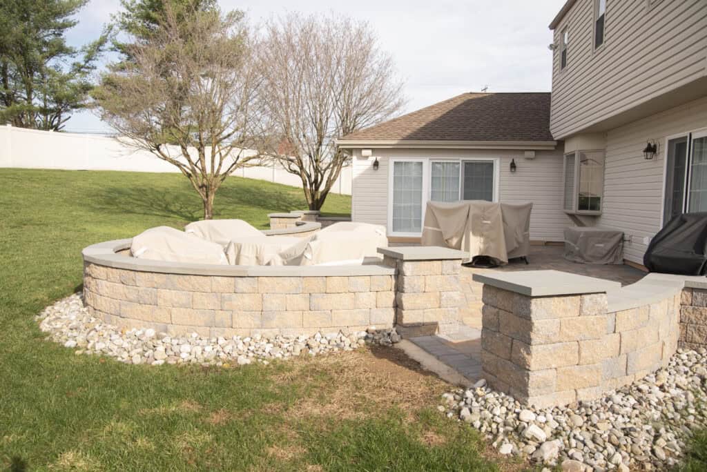 A backyard patio area with stone walls, covered patio furniture, and a barbecue grill next to a house. The patio is bordered by rocks and surrounded by grass and trees.