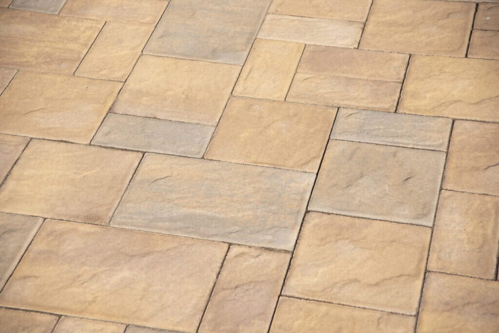 A close-up view of a paved surface made of rectangular and square beige and light brown stones arranged in an interlocking pattern.