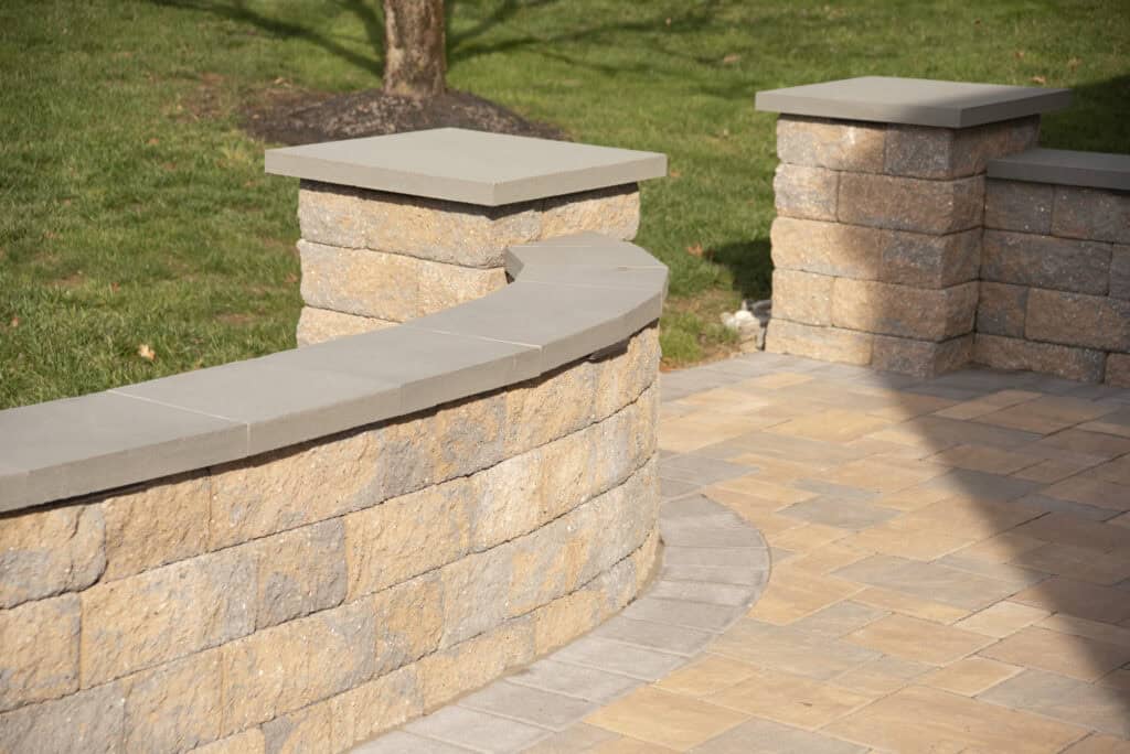 A curved stone retaining wall with a smooth concrete cap borders a paved patio area, with green grass and a tree visible in the background.