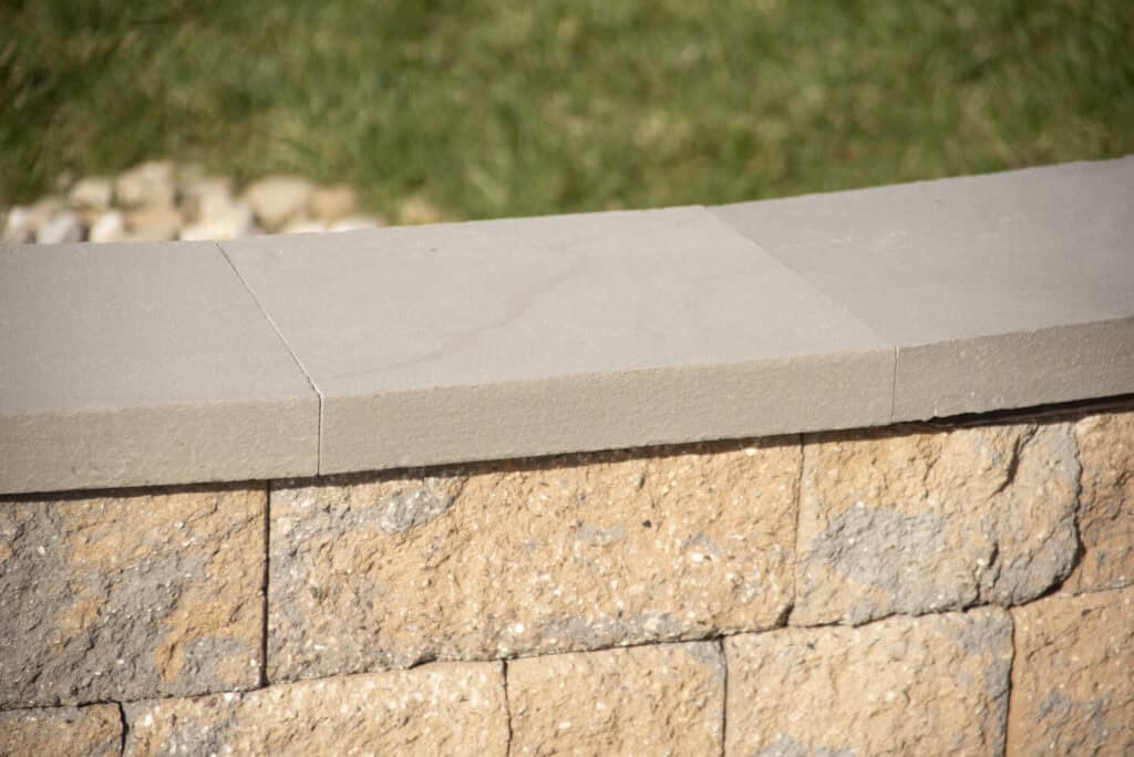 A curved stone retaining wall with smooth, light gray coping stones on top and rough, tan bricks below, set against a blurred background of green grass.