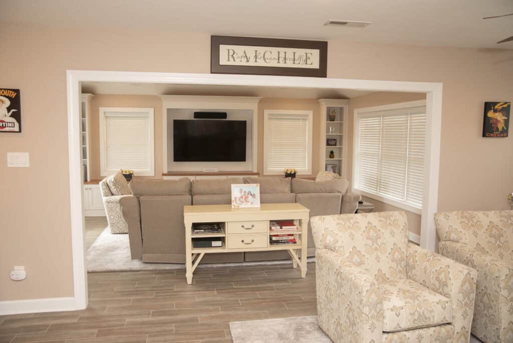 A cozy living room with beige walls, a large sectional sofa facing a wall-mounted TV, a console table with books and decor, patterned armchairs, and large windows with blinds letting in natural light.