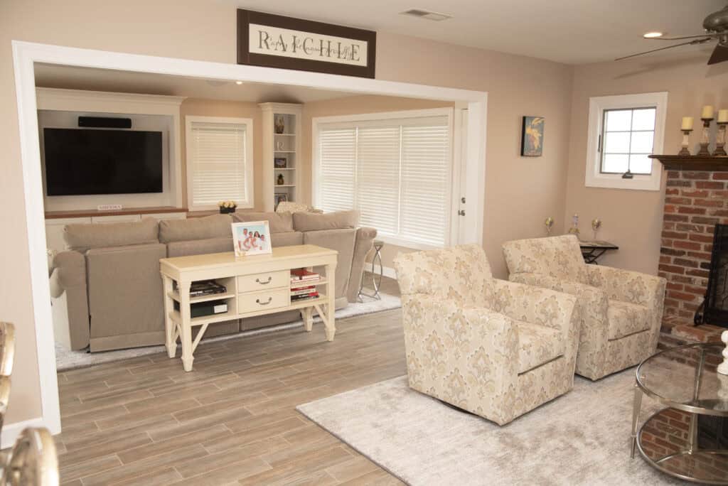 A cozy living room with beige walls and furniture, featuring two patterned armchairs, a white console table, a sectional sofa facing a TV, large windows with blinds, and a brick fireplace on the right.
