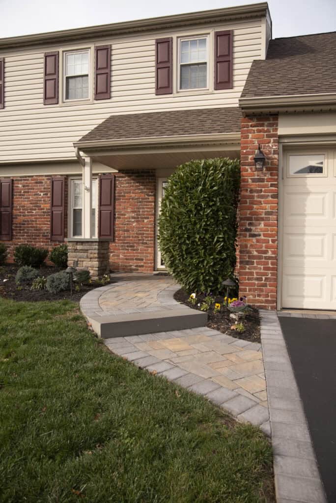 A paved walkway with steps leads to the front entrance of a brick and siding house with maroon shutters, a manicured lawn, and neatly trimmed bushes and flowers near the door.