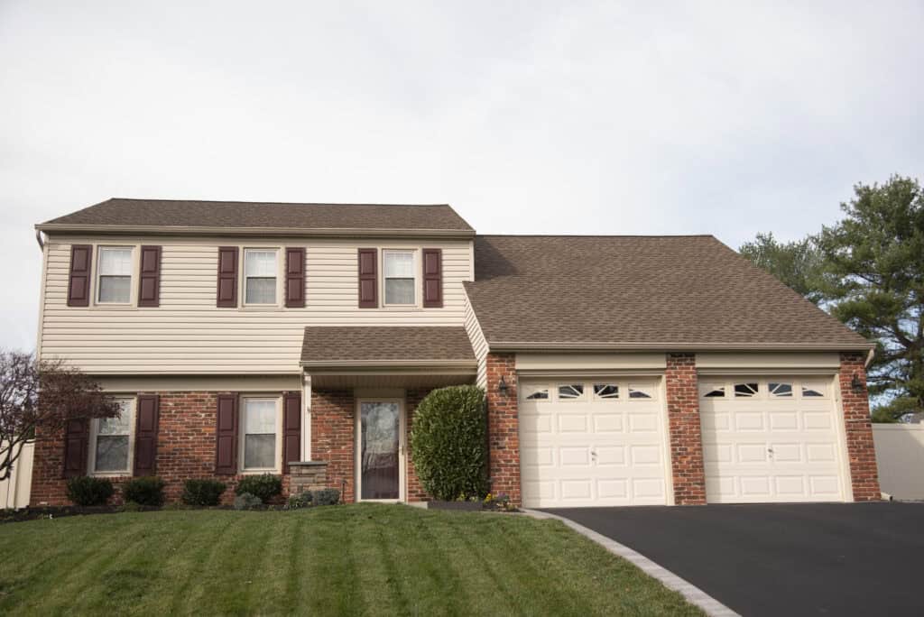 A two-story suburban house with beige siding, red brick accents, brown shutters, and a double garage. The front lawn is neatly trimmed, and a bush stands by the entrance. Pines and a fence are in the background.