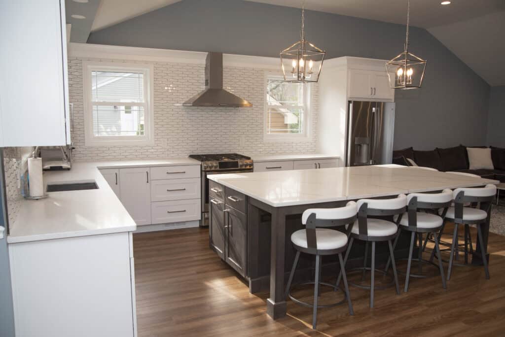 Modern kitchen with white cabinets, large island with four chairs, stainless steel appliances, wood flooring, geometric pendant lights, and white tile backsplash. Two windows let in natural light.