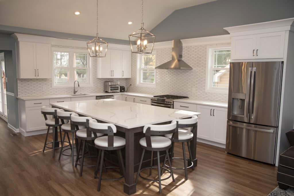 Modern kitchen with white cabinets, stainless steel appliances, a large island with a marble countertop, six bar stools, pendant lighting, wood flooring, and a tile backsplash. Natural light comes from multiple windows.