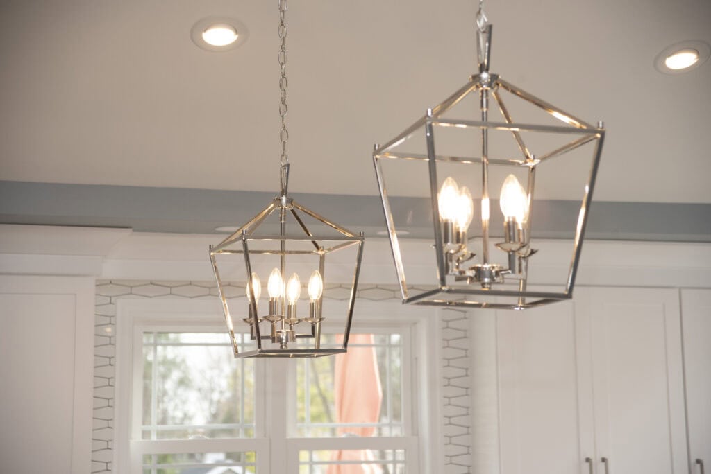 Two modern, geometric pendant light fixtures with exposed bulbs hang from the ceiling in a bright kitchen with white cabinets and a window in the background.