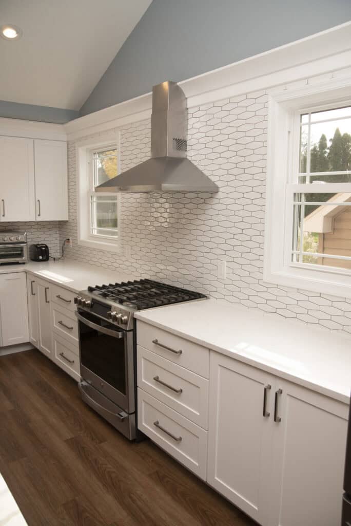 A modern kitchen with white cabinets, a stainless steel gas stove and hood, white hexagonal tile backsplash, and wood flooring. Two windows let in natural light above the countertop.