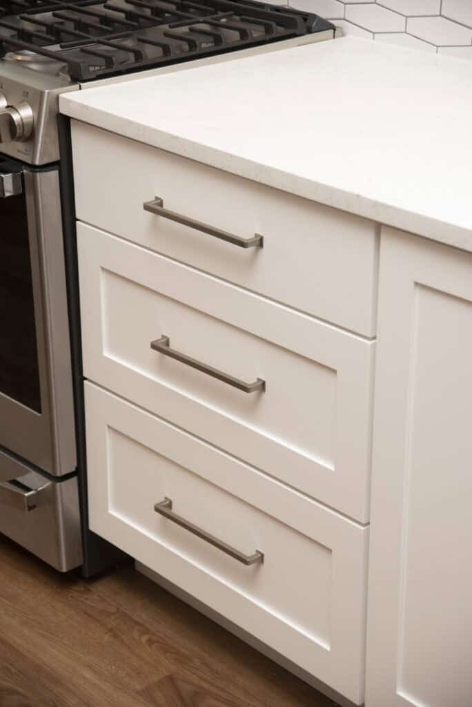 A close-up of a kitchen with white shaker-style drawers and silver handles next to a stainless steel gas stove. The countertop is white, and the floor is wood.