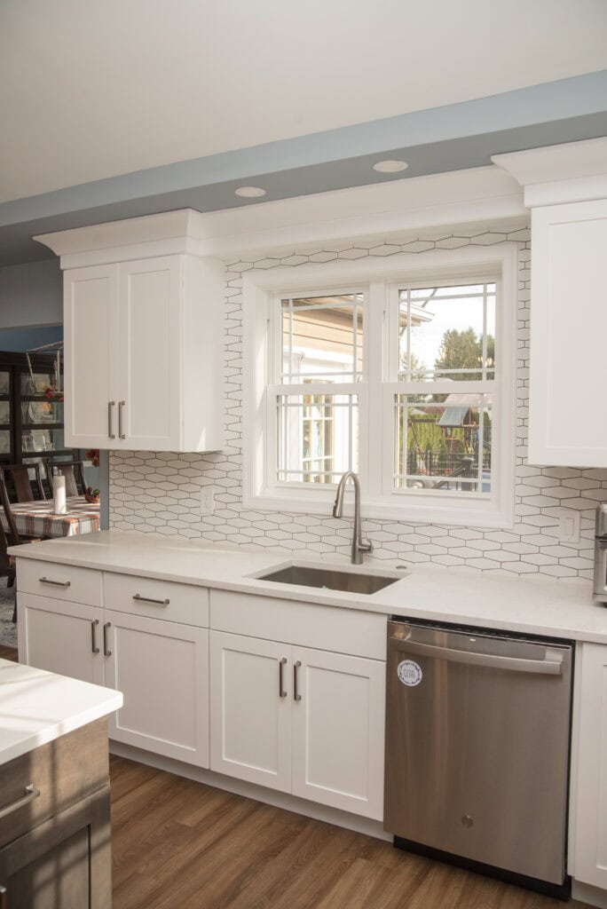 Bright, modern kitchen with white cabinets, a stainless steel sink under a large window, white subway tile backsplash, quartz countertops, and a dishwasher. Wood flooring adds warmth to the clean design.