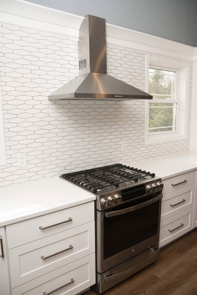 A modern kitchen with a stainless steel gas stove, matching range hood, white cabinets with metal handles, white countertops, and a white geometric tile backsplash. A window is visible beside the stove.