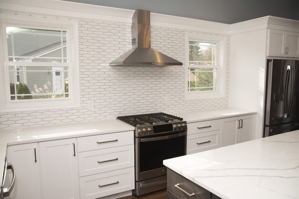 Modern kitchen with white cabinets, stainless steel appliances, a gas stove with a range hood, white hexagonal tile backsplash, marble countertops, and two windows providing natural light.