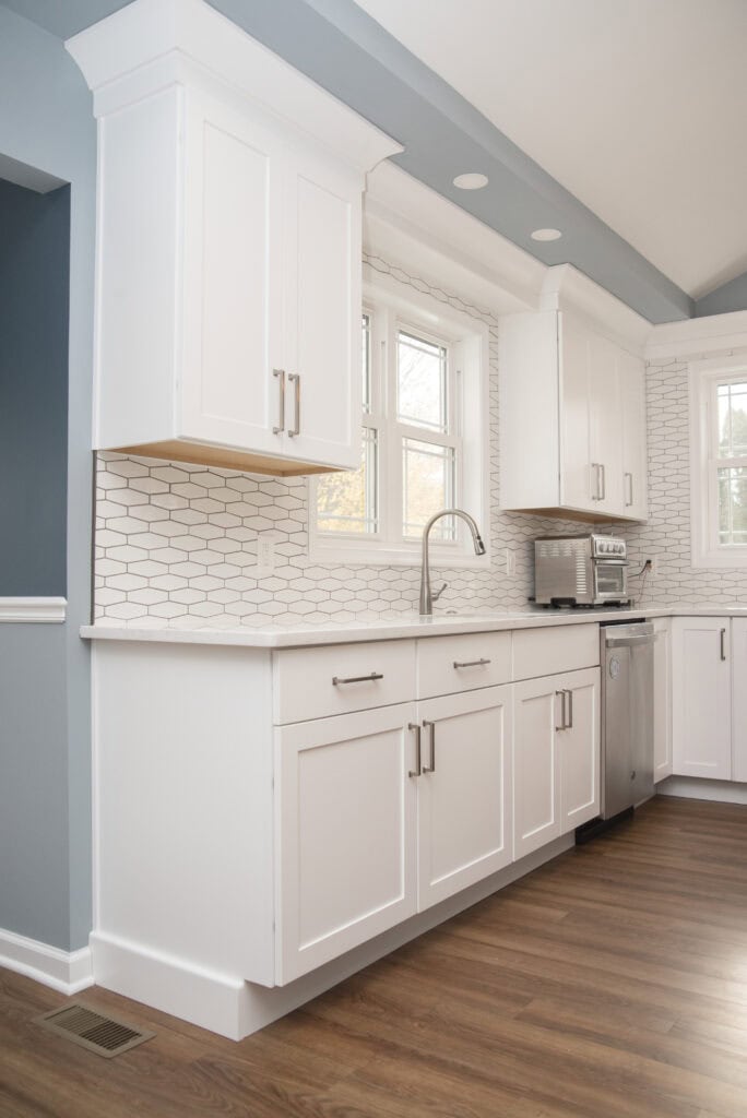 Modern kitchen with white cabinets, stainless steel appliances, hexagonal white tile backsplash, a large window above the sink, and wood flooring. The walls are painted a soft blue color.