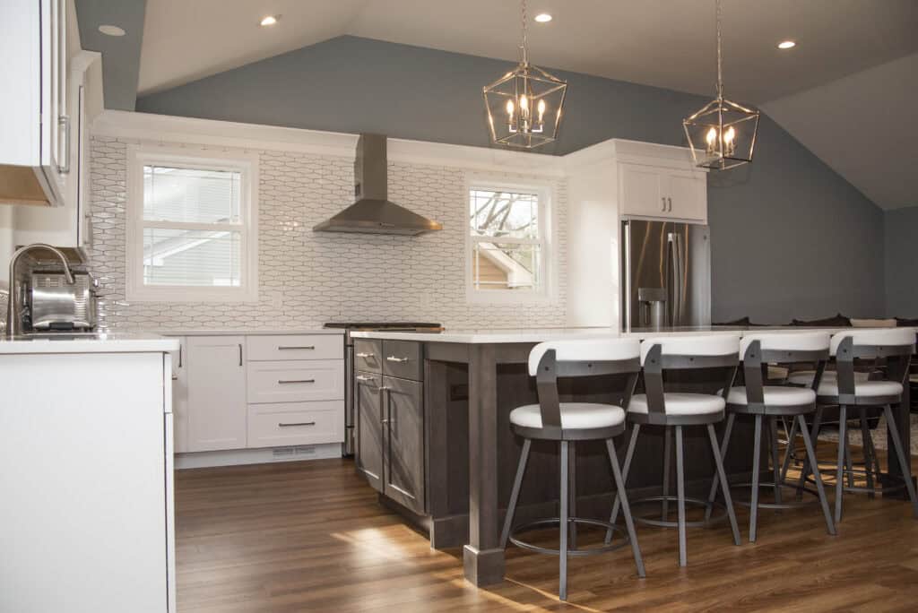 Modern kitchen with white cabinets, a large island with dark wood and white chairs, stainless steel appliances, geometric pendant lights, light wood floors, and a white tile backsplash. Natural light from two windows.