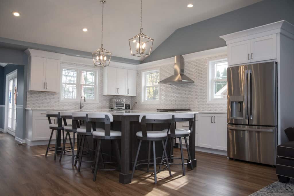 Modern kitchen with white cabinets, a large island with four stools, stainless steel appliances, pendant lights, hardwood floors, and a tile backsplash. Multiple windows provide natural light.