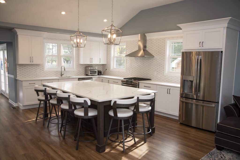 Modern kitchen with white cabinets, stainless steel appliances, a large marble island with six chairs, pendant lights above, wood flooring, and a tile backsplash. Natural light comes through multiple windows.