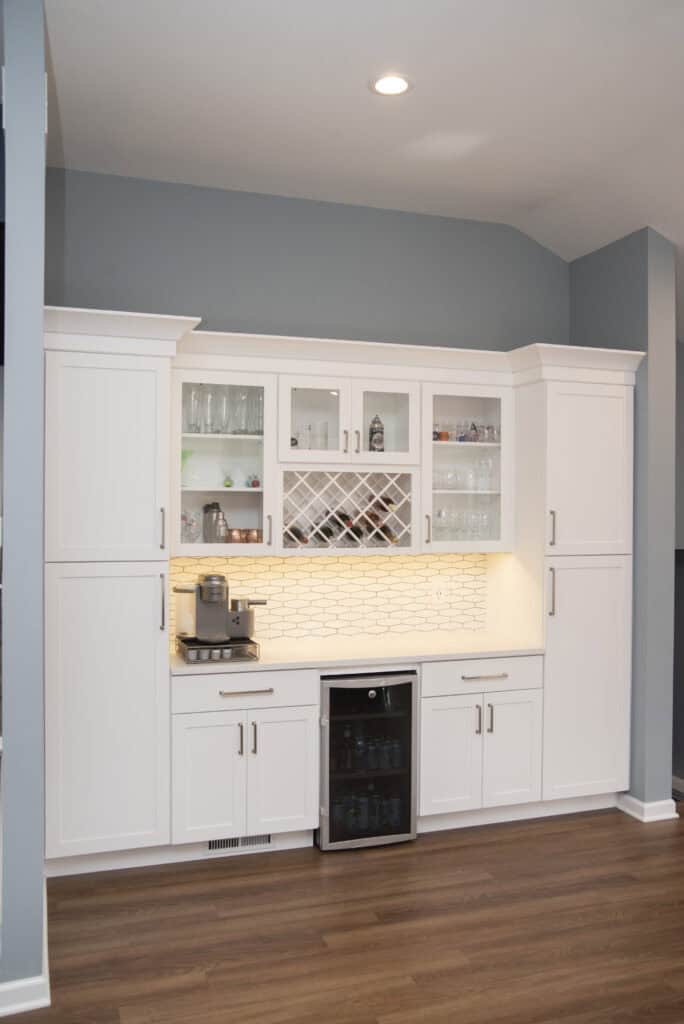 A modern white kitchen bar with glass cabinets, a built-in wine rack, beverage fridge, and coffee machine. The backsplash is white tile, and the space is accented by wood floors and soft overhead lighting.