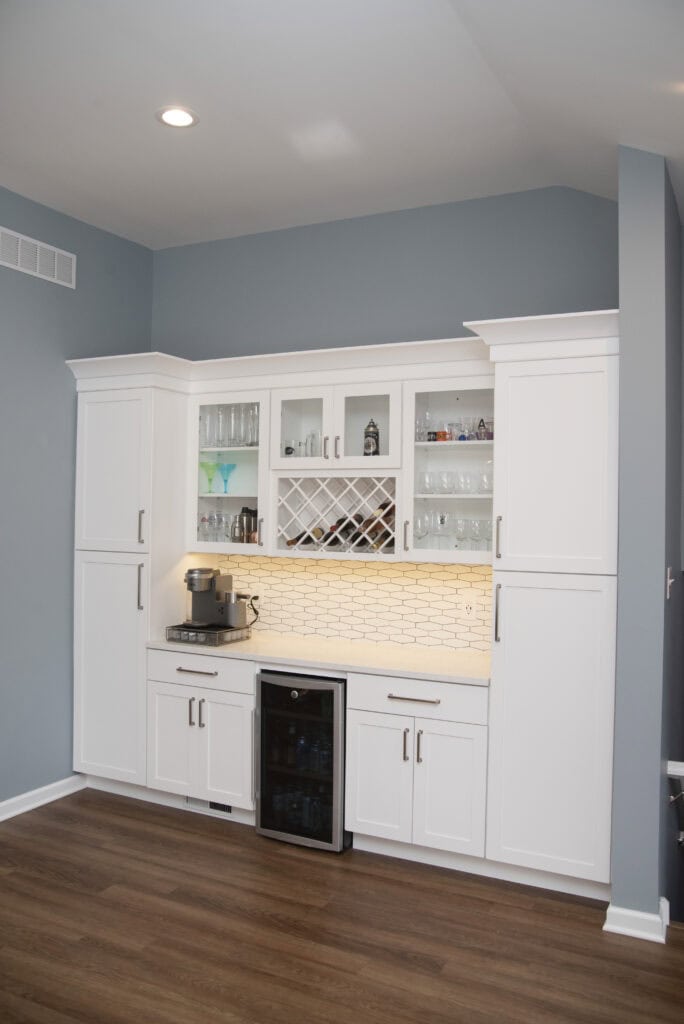 A modern home bar with white cabinets, glass shelves, a wine rack, under-cabinet lighting, a coffee maker, and a mini fridge, set against blue-gray walls and wood flooring.