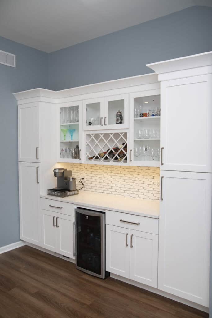 A modern home bar with white cabinets, glass-front upper cupboards, a wine rack, under-cabinet lighting, a coffee machine, and a built-in wine fridge against a blue wall and wood floor.