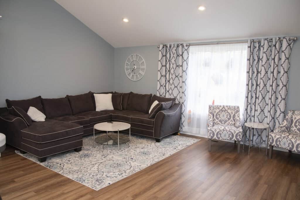 A cozy living room with a dark sectional sofa, white and gray patterned rug, round coffee table, two patterned chairs, light blue walls, wood flooring, and a large window with geometric curtains.