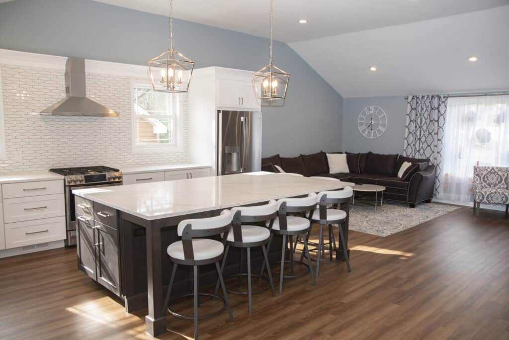 Modern open-concept kitchen and living room with brown wood floors, white cabinets, a large island with four chairs, stainless steel appliances, and a cozy seating area with a sofa and patterned curtains.