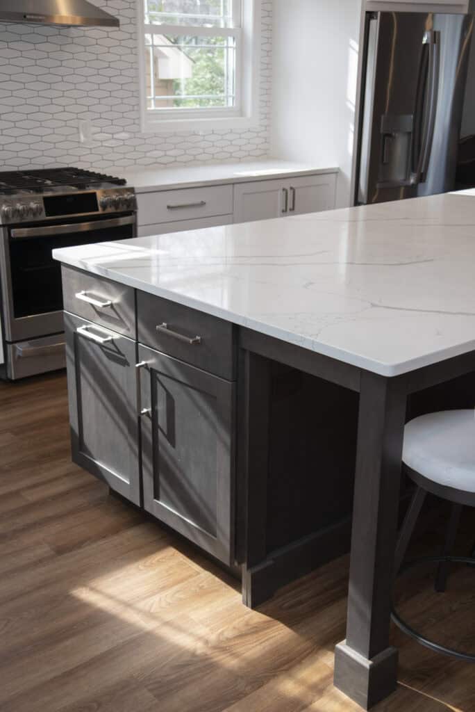 Modern kitchen with a large marble island, dark lower cabinets, white upper cabinets, stainless steel appliances, and wood flooring. Sunlight streams in through a window above the counter.