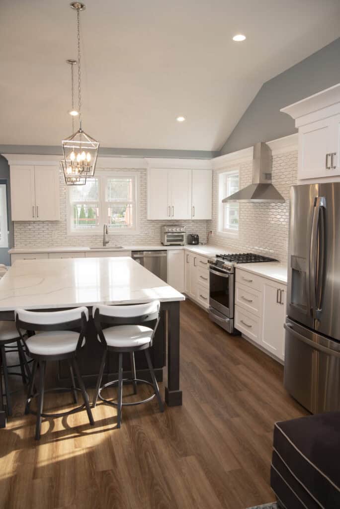 Modern kitchen with white cabinets, stainless steel appliances, a large island with white countertops, four chairs, wood flooring, pendant lighting, and windows letting in natural light.