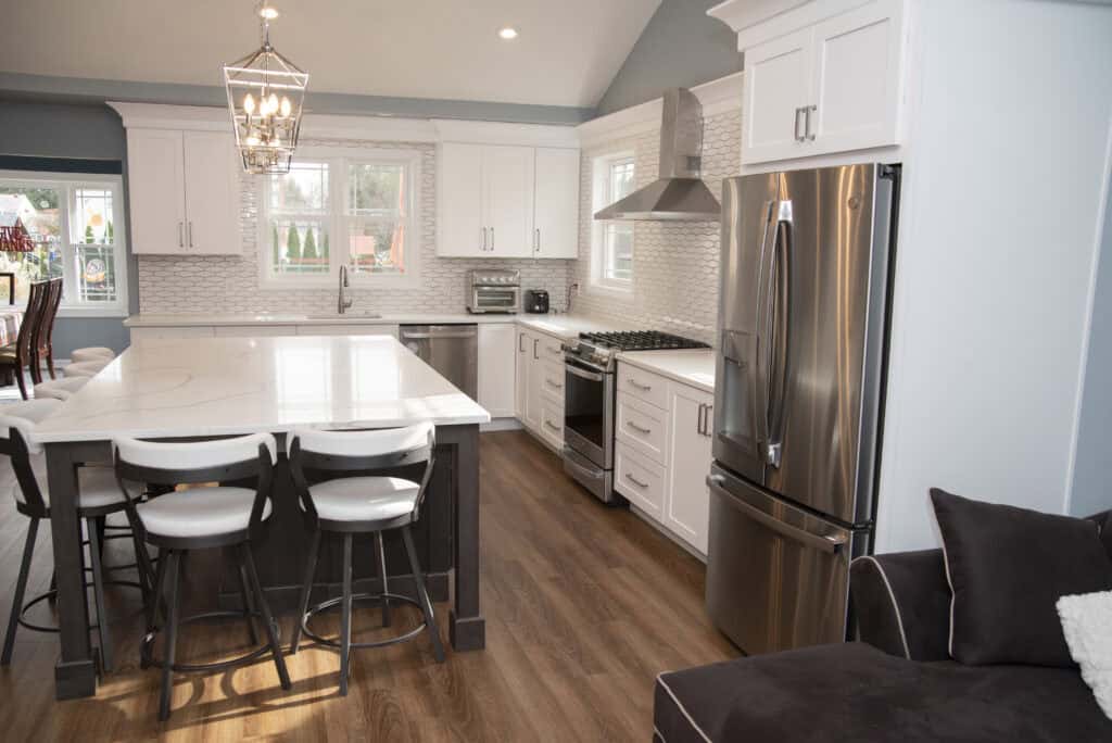 Modern kitchen with white cabinets, stainless steel appliances, marble island with four chairs, wood flooring, pendant light, and a gray sofa in the foreground. Large windows provide natural light.