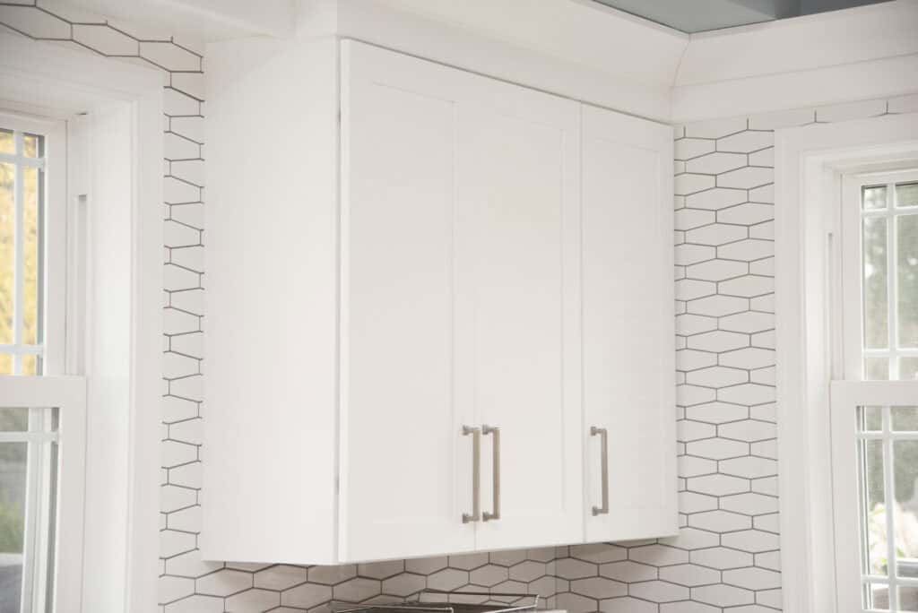 White kitchen wall cabinet with three doors and silver handles, mounted between two windows. The backsplash features white hexagonal tiles with gray grout. Natural light is coming in through the windows.
