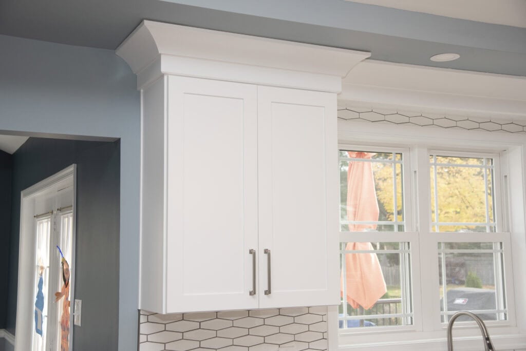 White kitchen cabinets with silver handles are mounted on a blue wall above a backsplash with hexagonal tiles. Large windows with white frames and an orange umbrella outside are visible.