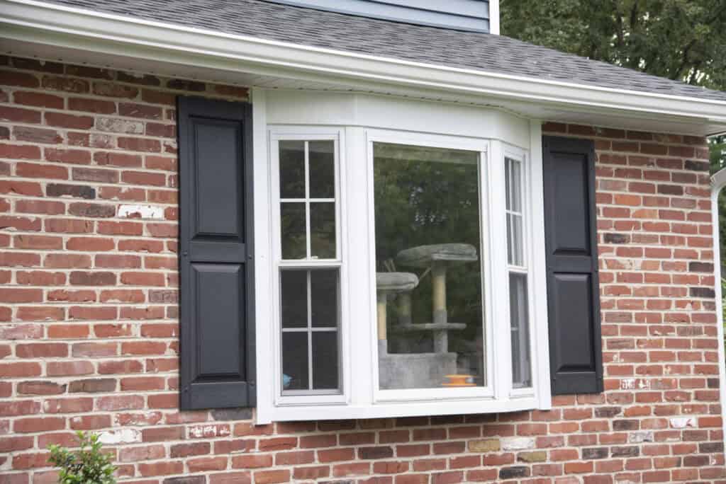 A brick house with a bay window featuring black shutters; inside the window, a cat tree is visible, and greenery and trees are reflected in the glass.