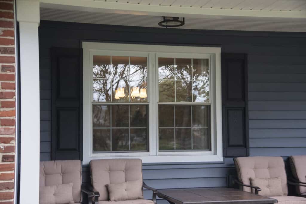 A double window with white trim and black shutters is set in a blue house exterior. Two cushioned chairs and a table sit in front of the window on a porch with a white ceiling. Trees reflect in the window glass.