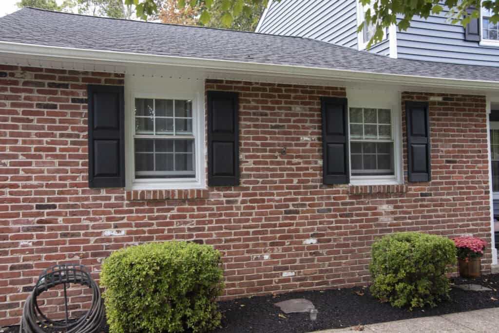 A red brick house with two white-trimmed windows featuring black shutters. Small green bushes and a coil of garden hose are in front, and pink flowers sit near the sidewalk.