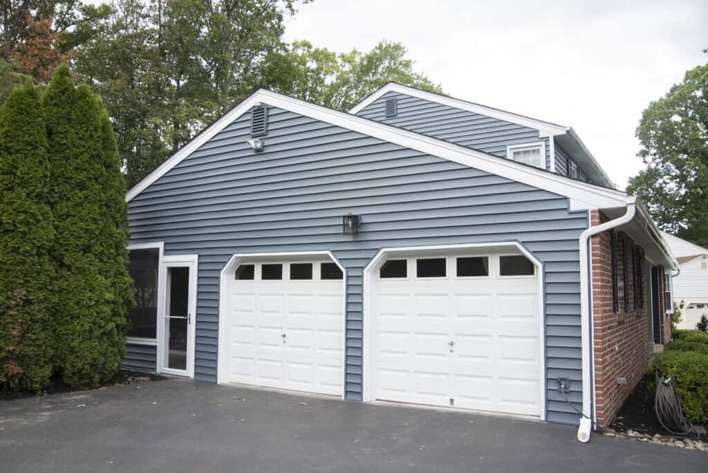 Blue house with white trim, featuring a double garage with two white doors, a paved driveway, and green trees in the background.